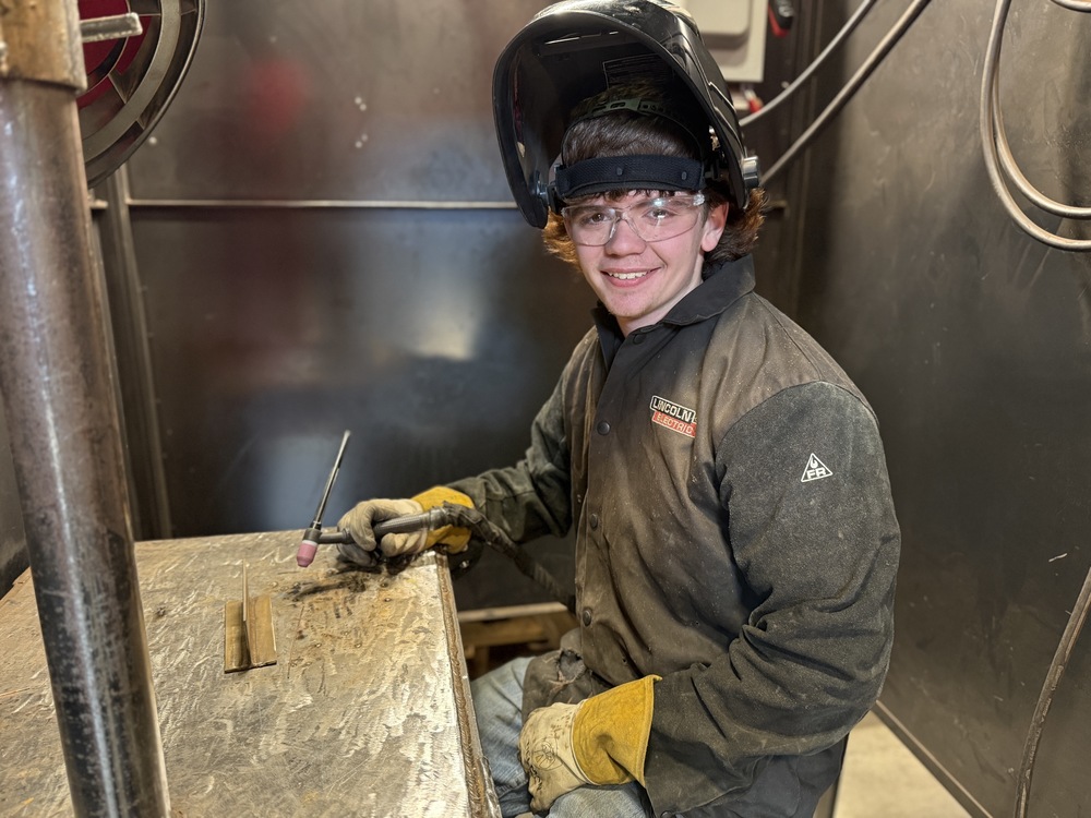 Student dressed in welding jacket and helmet sits at a table