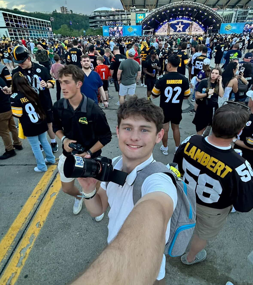 Young man takes a selfie with a camera in hand amid a large crowd in football jerseys at an outdoor stadium event with a stage and cloudy sky in the background.