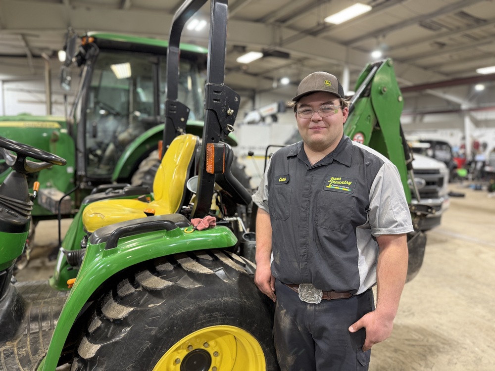 A student in a John Deere uniform stands beside a green tractor in a workshop, with additional equipment visible in the background.