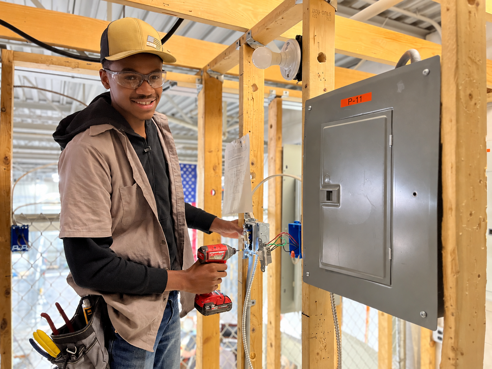 Electrical Technology student wearing safety glasses smiles toward the camera while using a cordless drill to work on an electrical box mounted beside a breaker panel in a wooden training structure.