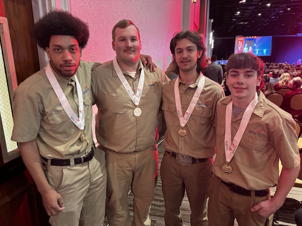 Four BCTC students wearing tan SkillsUSA uniforms stand together at the SkillsUSA Pennsylvania awards ceremony in Hershey, each wearing a medal around their neck. The students smile for a photo near the auditorium entrance, with the stage and audience visible in the background.