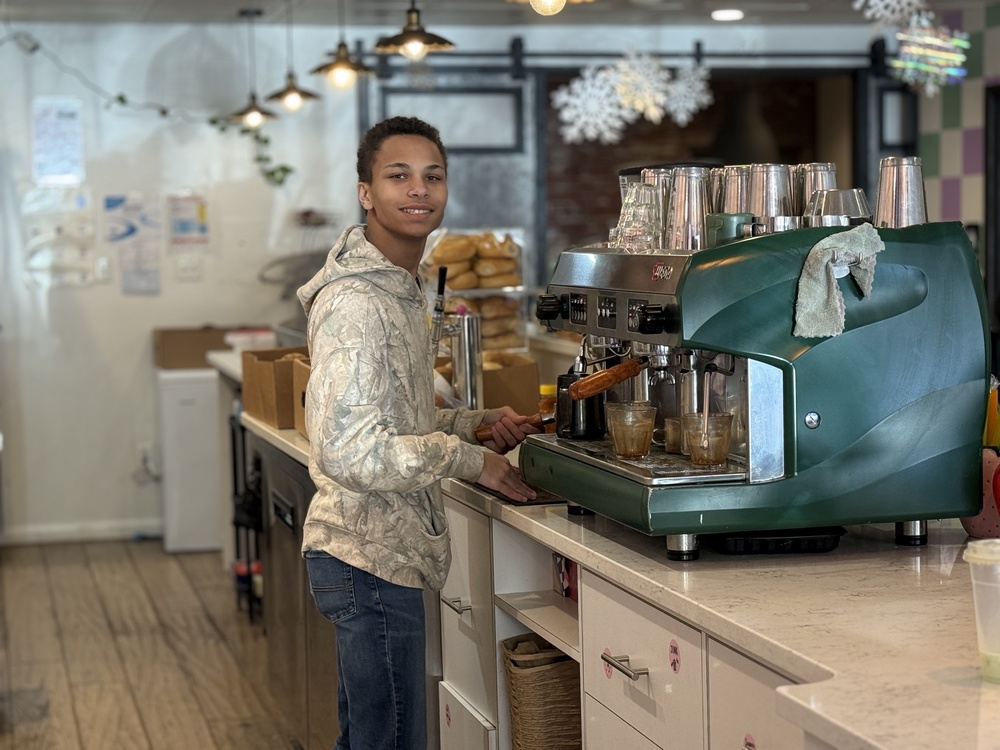 Student stands behind a café counter using an espresso machine, preparing drinks with cups and equipment arranged on the workspace.