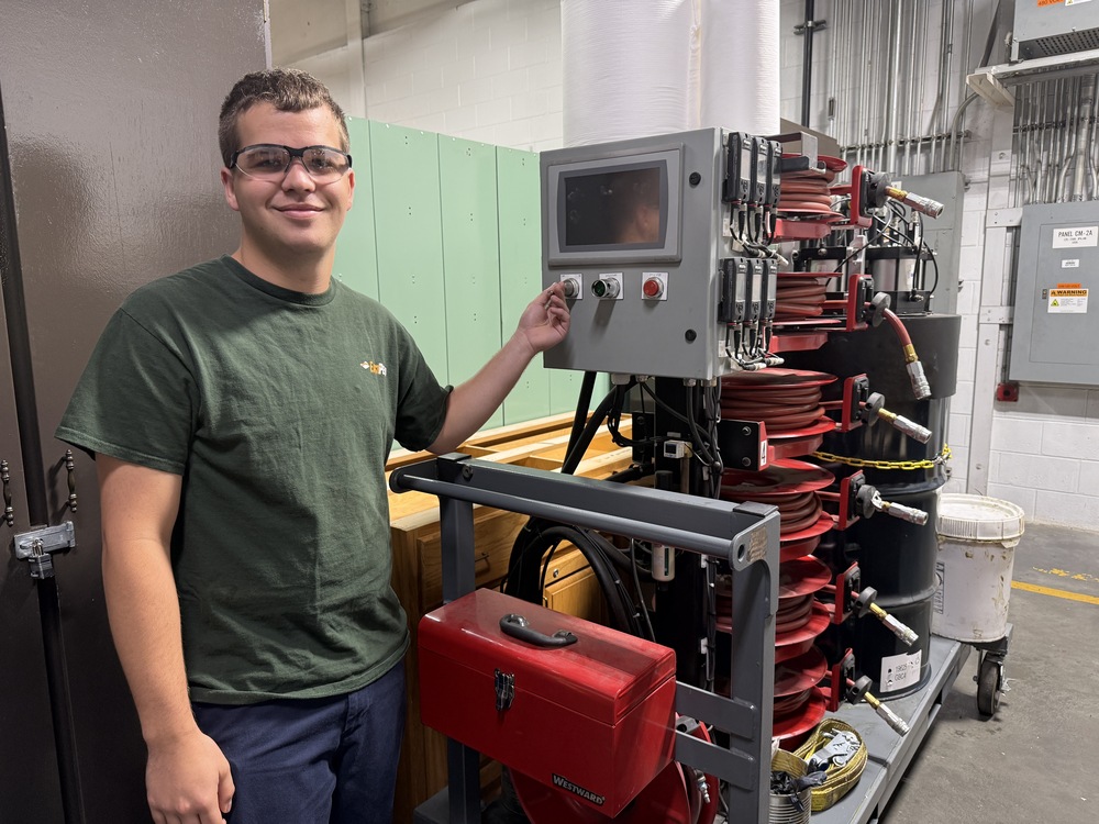Student wearing safety glasses stands beside industrial equipment, holding a control switch on a panel attached to a mobile hose reel system.