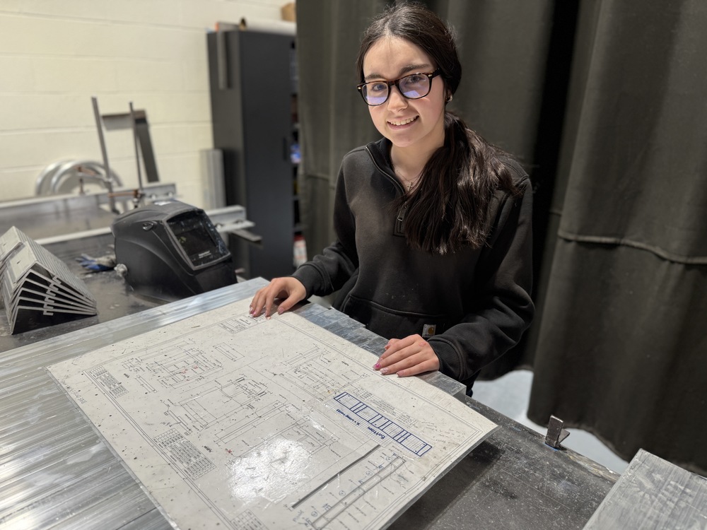 Student in a fabrication lab reviews a technical blueprint on a metal worktable, with welding equipment and shop tools nearby.