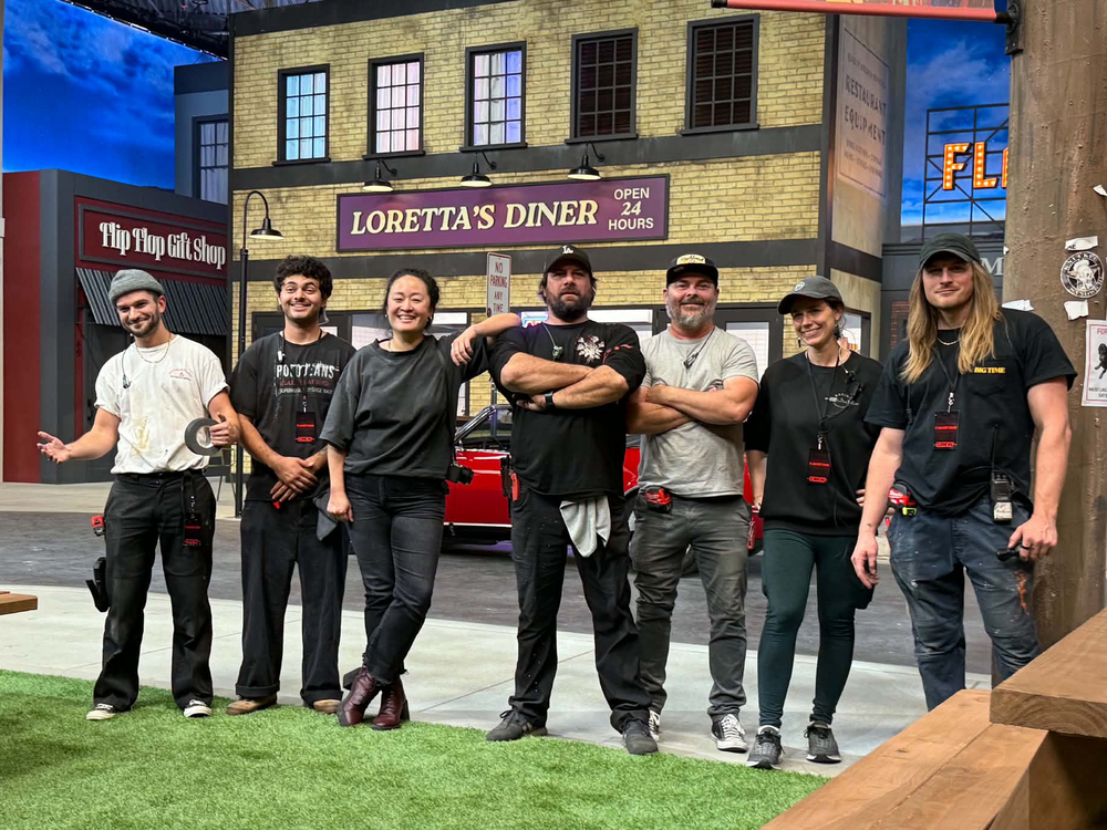 Seven production crew members pose together on a colorful television set in front of a brick storefront labeled “Loretta’s Diner.”