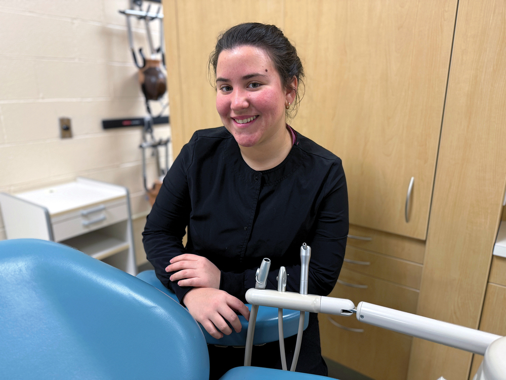 Student in a clinical setting smiles beside a dental chair, with dental tools and equipment visible in the foreground and cabinets and medical training equipment in the background.