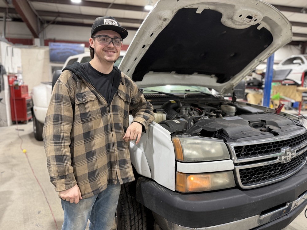 Student wearing safety glasses and a cap smiles while standing beside a vehicle with the hood open in an automotive shop, with the engine visible.