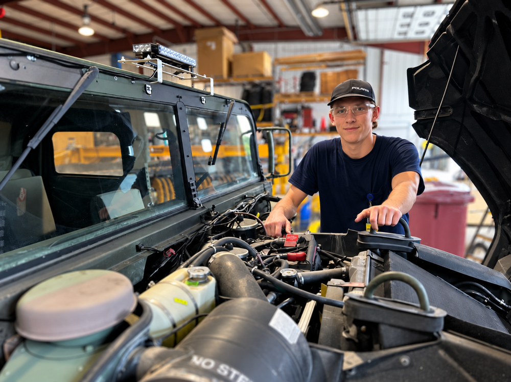 A young man wearing a cap and clear safety glasses stands in an automotive workshop, leaning over the open engine bay of a large utility vehicle while looking toward the camera, with the vehicle’s hood raised and tools and shop equipment visible in the background.