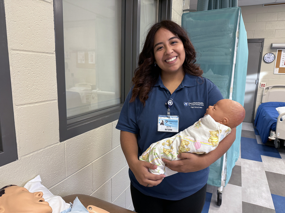 A high school intern wearing a navy Penn State Health polo stands in a healthcare lab, smiling while holding a swaddled infant training mannequin. Behind her are hospital beds, medical curtains, and classroom equipment. A second mannequin is partially visible on a nearby bed.