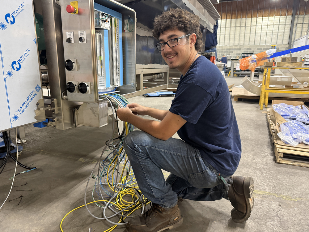 Student wearing safety glasses kneels beside industrial equipment while working with a bundle of electrical wires connected to an open control panel.