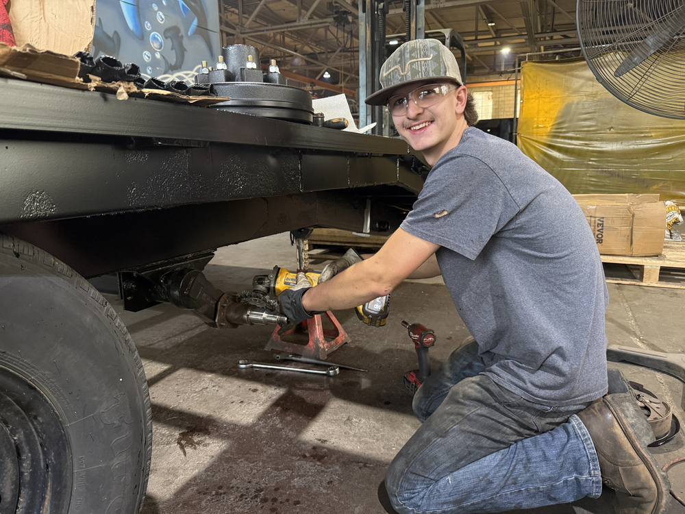 Student wearing safety glasses and work gloves kneels beside a raised trailer while using a power tool to work on the wheel axle assembly.