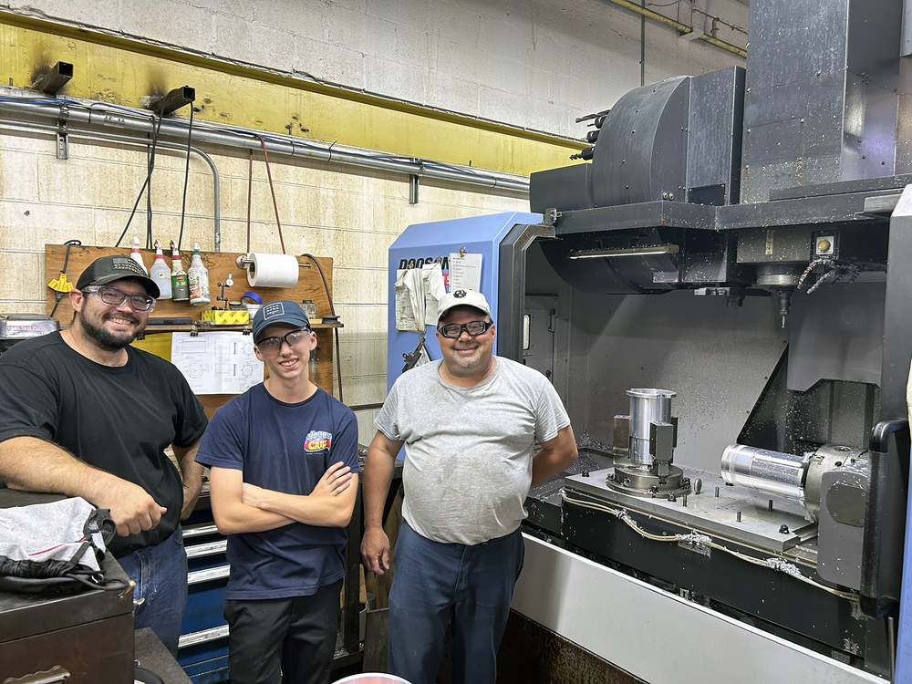 Three people stand smiling in a machine shop next to a large CNC machine. All three wear safety glasses; two wear baseball caps. Metal parts and machining equipment are visible on the workbench and inside the CNC machine, with tools, drawings, and supplies hanging on the wall behind them.