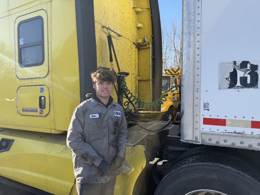 A student in a transportation training program stands between a yellow semi-truck cab and trailer, wearing work gloves and a coverall, with air lines and truck components visible behind him in an outdoor work area.