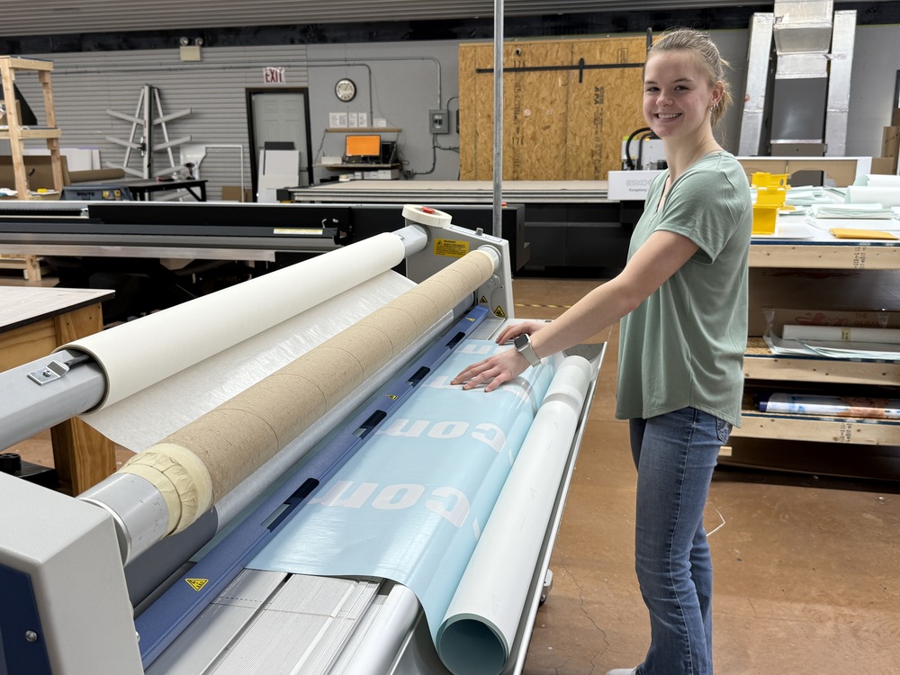 A student smiles while guiding blue material through a large-format laminator in a career and technical education lab.