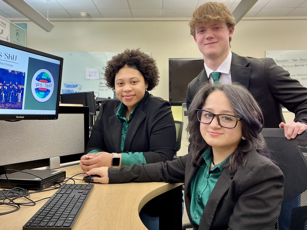 Three Business Management and Entrepreneurship students in professional attire pose at a computer in a classroom, with the Res-SKU logo displayed on the monitor.