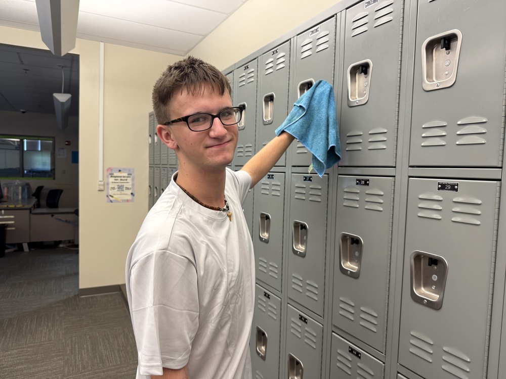 Student wearing glasses and a white shirt wipes down a row of gray lockers with a blue cloth inside a BCTC classroom.