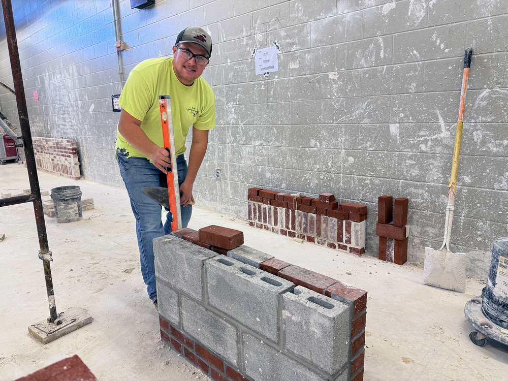 A student in a construction training lab uses a level to check the alignment of a partially built masonry wall made of concrete blocks and bricks, with tools and stacked bricks visible in the background.