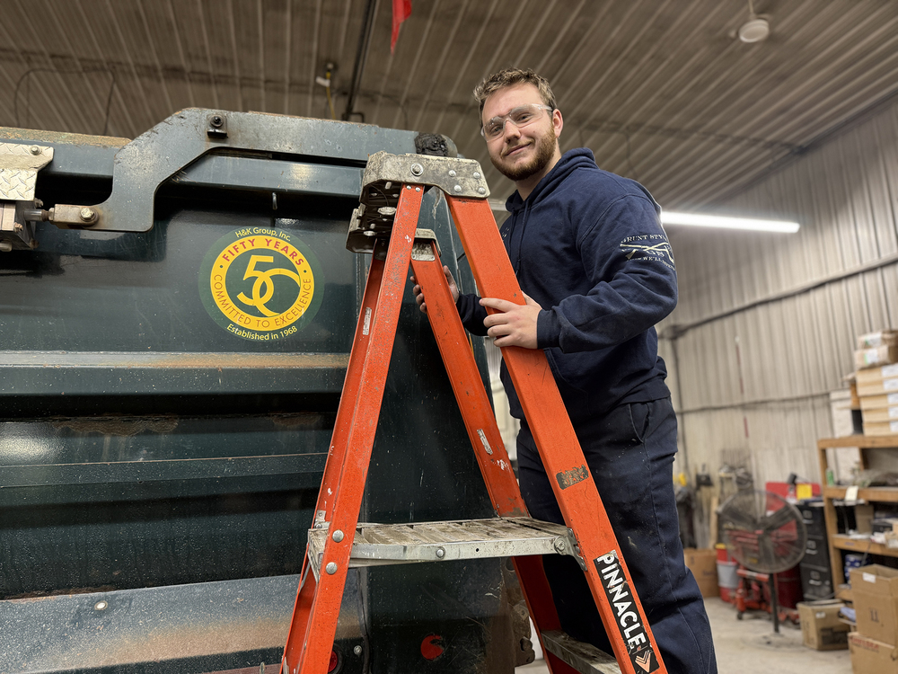 Student wearing safety glasses stands on a ladder next to a construction vehicle inside a workshop.