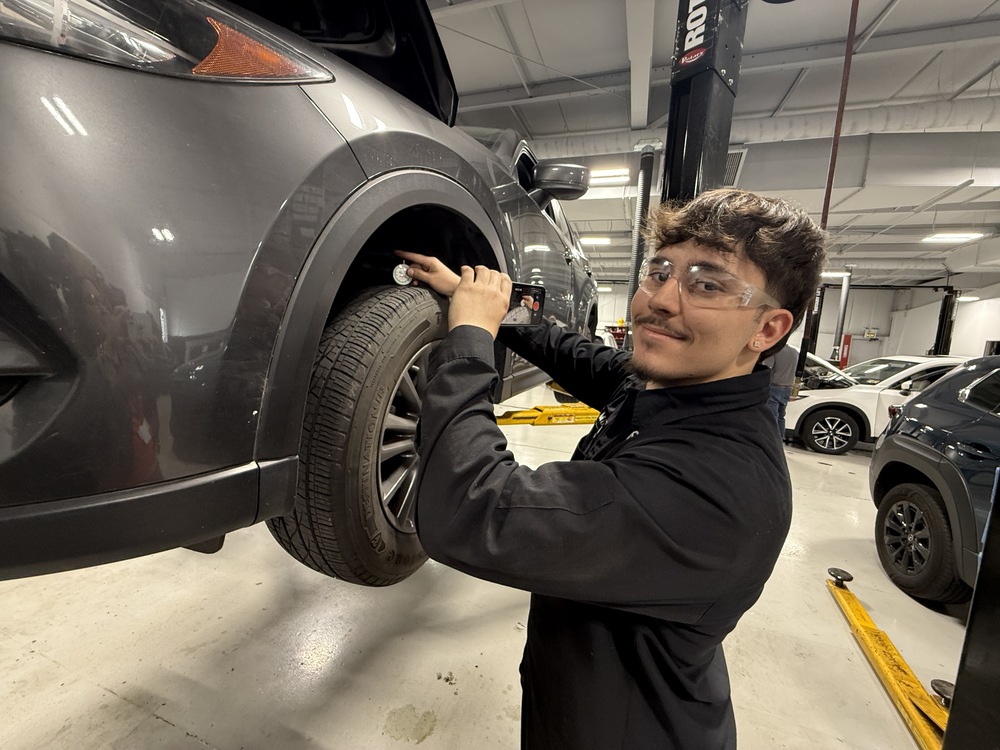 A student wearing safety glasses checks the wheel area of a vehicle raised on a lift in an automotive lab.