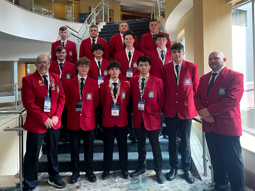 A group of thirteen individuals stands on a staircase inside a building. They are all wearing red blazers, white shirts, black ties, and black pants. Each person has a name badge hanging from their neck. The background shows part of the building's interior with glass railings and windows.