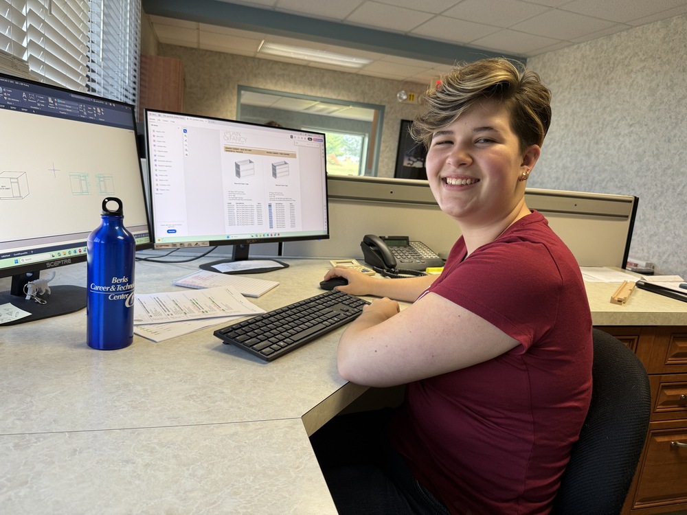 Student smiles at the camera while seated at a workstation with two computer monitors displaying technical drawings and product information.