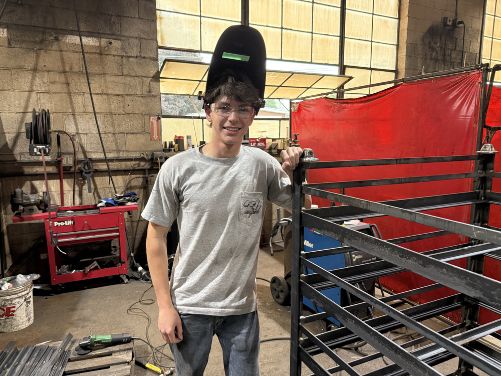 Student wearing safety glasses and a raised welding helmet stands beside a metal frame in a welding shop.