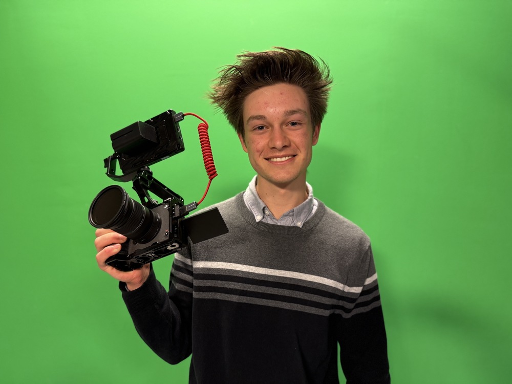 A person stands in front of a green screen, holding a professional video camera rig. The camera rig includes a large lens, an external monitor, and a red coiled cable connecting components. The person's face is pixelated for anonymity. They are wearing a gray sweater with white and dark gray stripes over a light blue collared shirt.