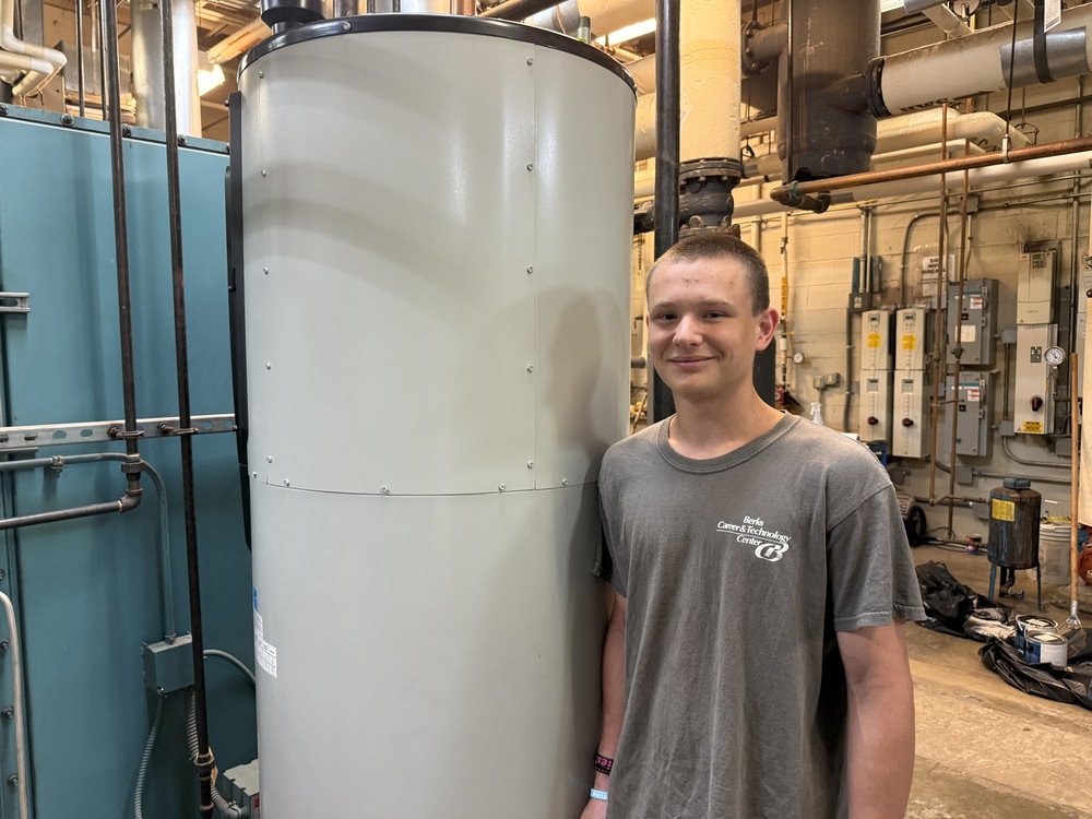 A BCTC student smiles while standing beside a large cylindrical HVAC unit in an industrial mechanical room with pipes and equipment in the background.