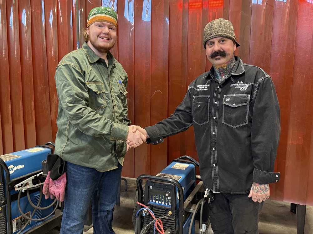 A welding student and instructor stand side by side in a workshop, shaking hands in front of welding equipment. The student wears a green protective jacket, gloves, and a welding cap, while the instructor wears a black work shirt with "Berks Career & Technology Center" embroidered on it and a patterned beanie. Both are smiling, with a red safety curtain in the background.