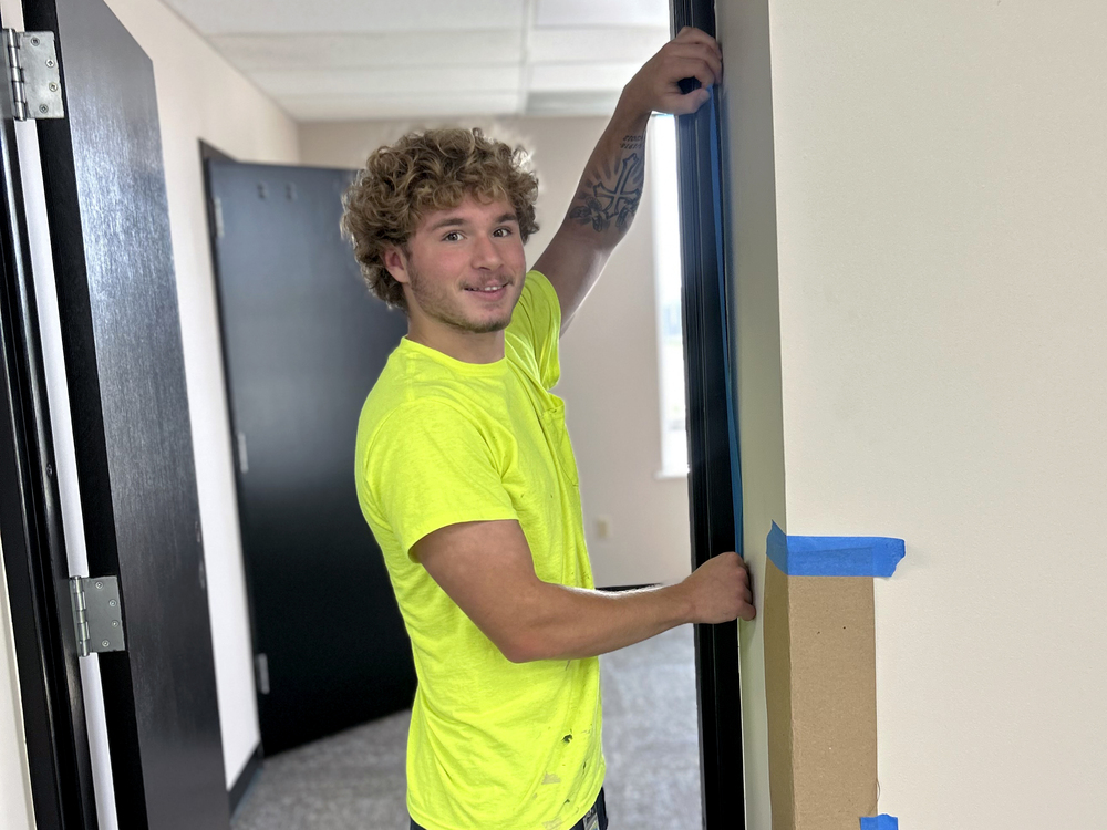 Student wearing a bright yellow shirt smiles toward the camera while positioning black door trim along a doorway in an interior hallway.