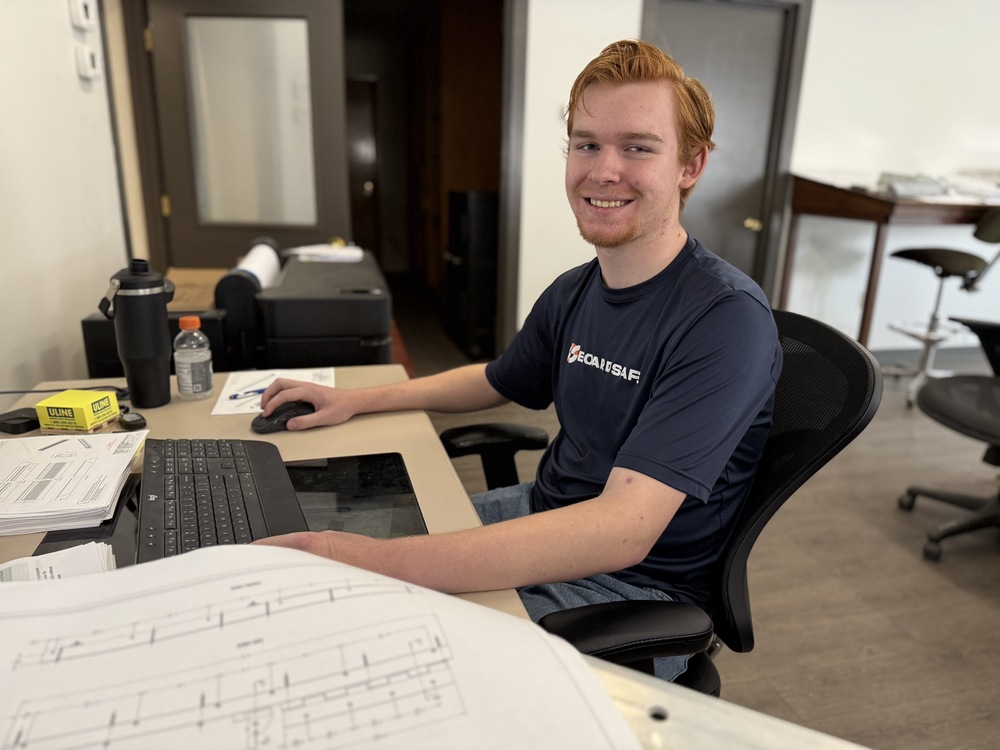 A student smiles while seated at a computer workstation with printed technical drawings spread across the desk.