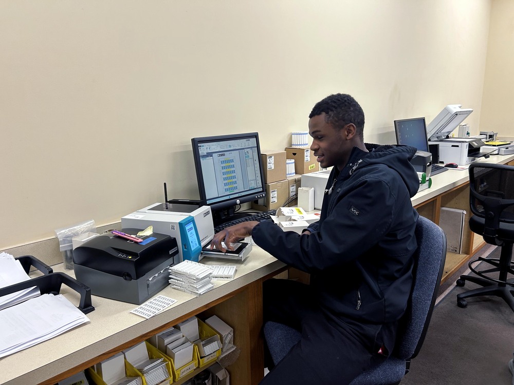 A student works at a labeling and printing workstation, using equipment beside a computer monitor with stacks of labels and supplies arranged across the counter.