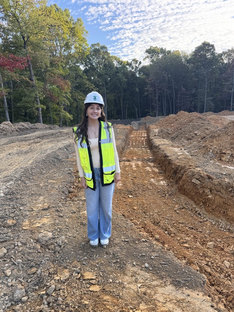 A young woman stands on a construction site wearing a light blue hard hat and a bright yellow reflective safety vest. She is smiling, dressed in a beige long-sleeve shirt and light blue jeans, with trees and a partly cloudy sky in the background. The ground around her shows freshly dug trenches and piles of dirt, suggesting active site work.