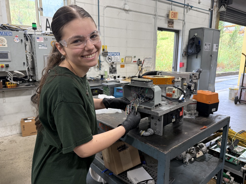 Student wearing safety glasses and gloves smiles while working with exposed wiring on a piece of industrial equipment in a technical lab.