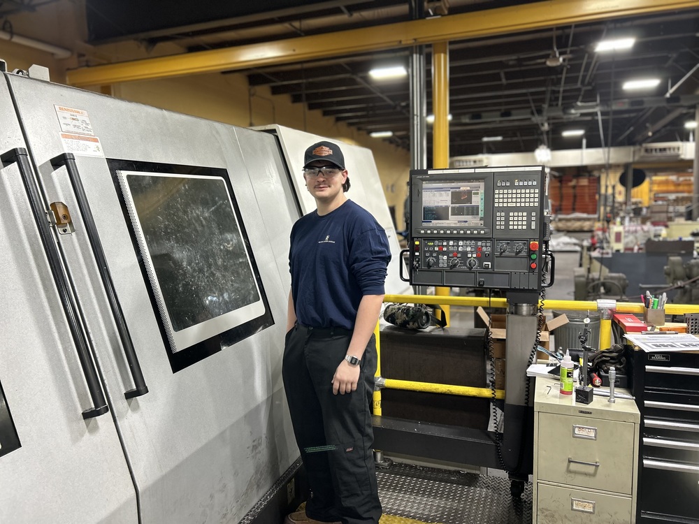 Student wearing safety glasses stands beside a CNC machine and control panel inside a manufacturing lab.