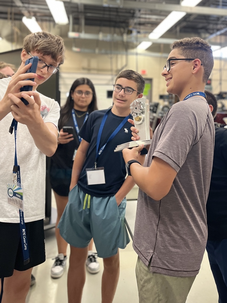 a group of people indoors, likely in a workshop or classroom setting. One person is holding a device or model, and another is taking a photo of it with a smartphone. The participants are wearing name tags and lanyards, indicating they might be part of an organized event or group activity.