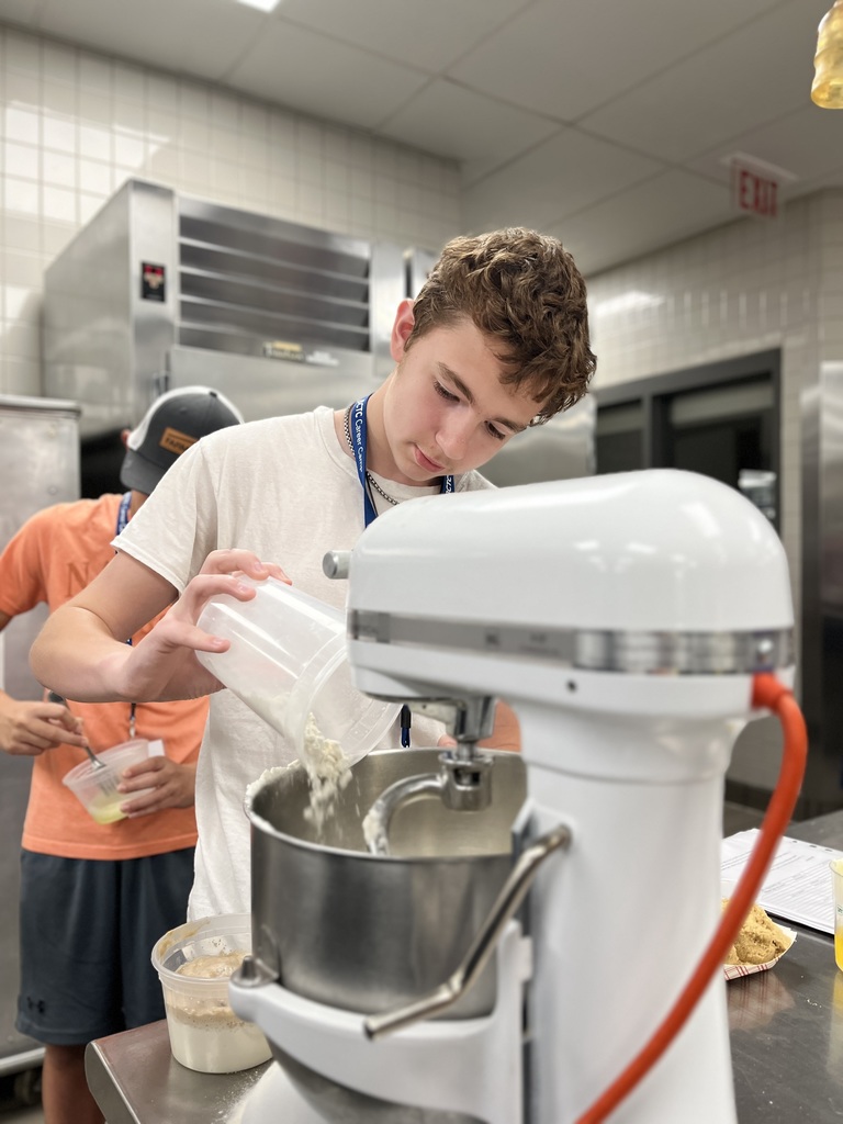 a person in a kitchen pouring an ingredient from a container into a stand mixer. Another individual in the background is also holding a container with ingredients. The kitchen features stainless steel appliances and tiled walls, suggesting a professional or educational culinary environment.