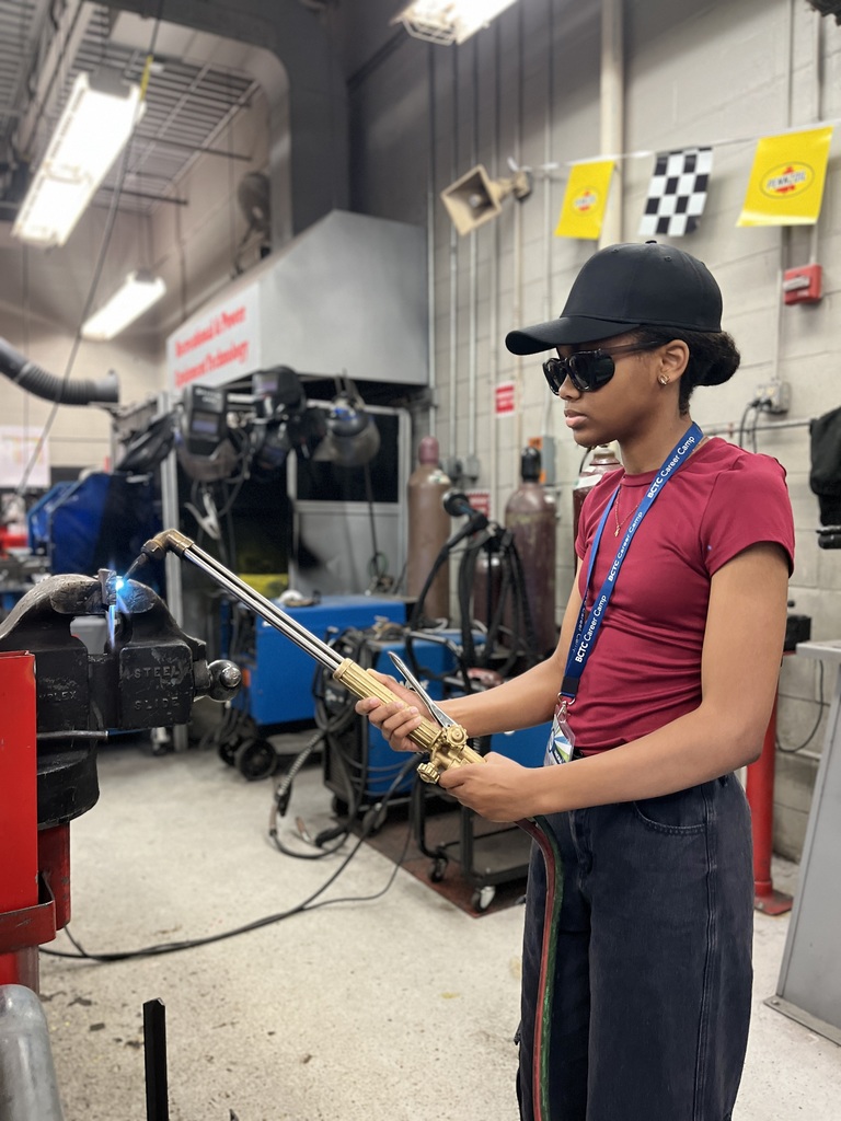 A person wearing a red shirt, dark pants, and a black cap is using an oxy-acetylene torch in a workshop. The person is holding the torch with both hands, directing the flame towards a metal object clamped in a vise. The workshop has various equipment and tools in the background, including gas cylinders and machinery.