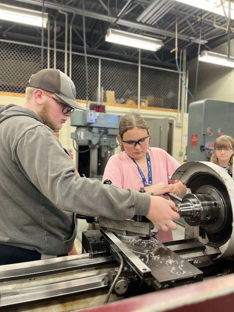three individuals in a workshop environment, working with a metal lathe machine. One person is wearing a gray hoodie and cap, another is in a pink shirt with a lanyard, and the third is only partially visible. The background includes additional machinery and storage shelves, indicating a hands-on technical or vocational setting.