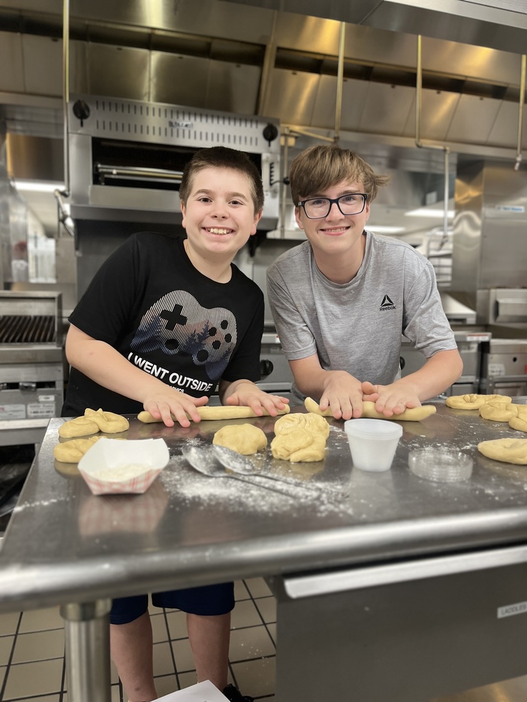 two individuals working in a kitchen, rolling out dough on a stainless steel table. There are multiple pieces of dough and some flour visible on the table surface. In the background, you can see kitchen appliances like ovens and grills, indicating a culinary or baking activity.
