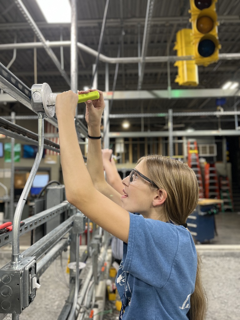 The image shows a person engaged in electrical work, using a tool to adjust or install components on a metal framework. The setting appears industrial, with visible elements like ladders, cables, and a traffic light in the background.