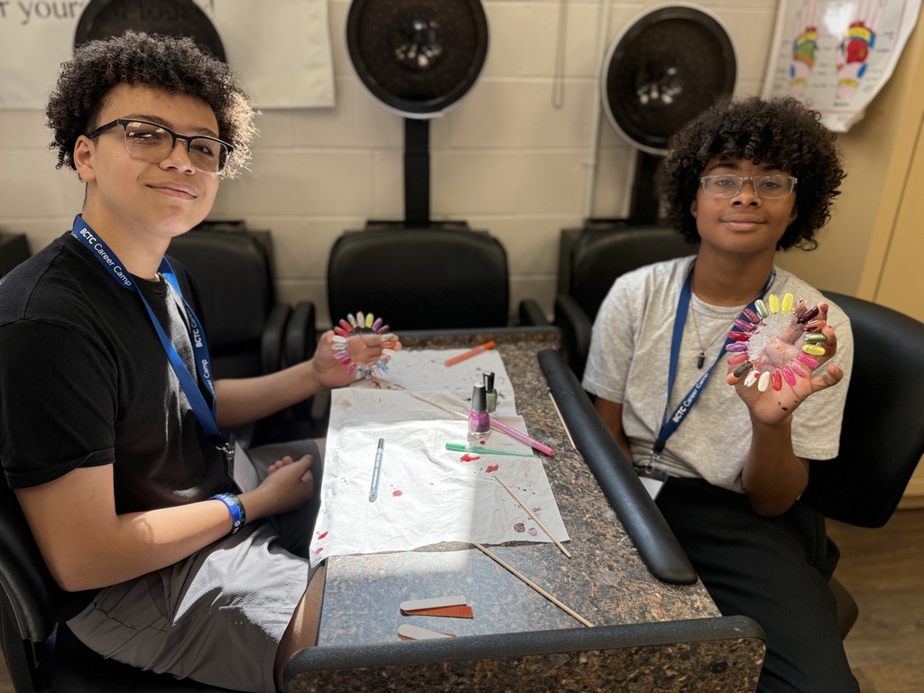 A person with curly hair sits on a black salon chair, wearing a black t-shirt, gray shorts, and a blue lanyard. They hold a circular nail polish color palette in their left hand. On the table in front of them are two white paper towels with pink dots, an open bottle of purple nail polish, and several colored markers or pens. Two black hair dryers are mounted on the wall behind them.