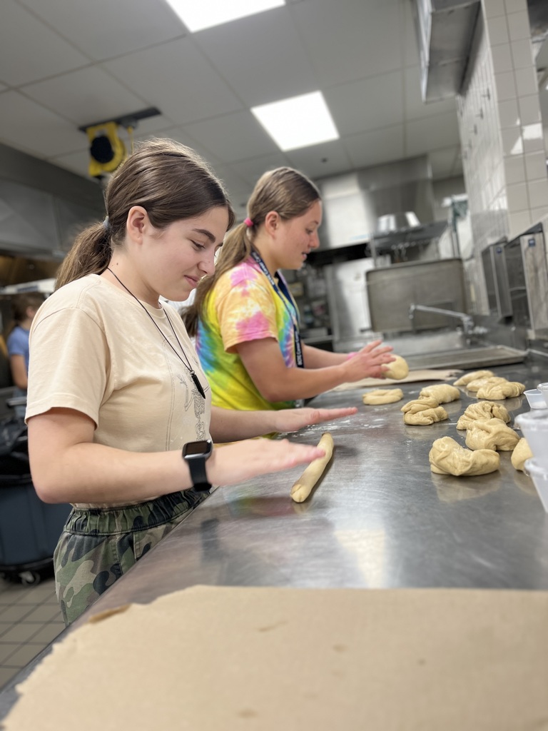 Two individuals are working with dough in a commercial kitchen. One person in the foreground is rolling dough into a long, thin shape on a stainless steel countertop, while the other in the background is shaping dough into knots or rolls. Several pieces of dough, both shaped and unshaped, are visible on the counter. The kitchen features stainless steel appliances and tiled walls.