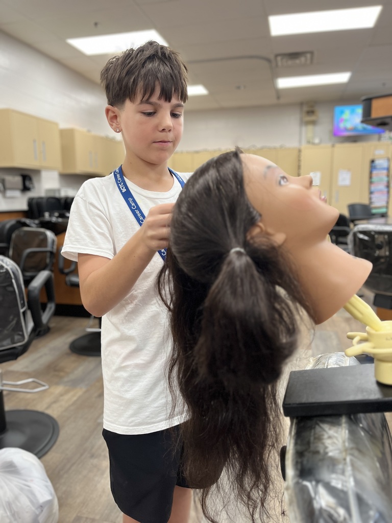 A person is standing in a classroom or training room, working on the hair of a mannequin head with long dark hair. The individual is wearing a white shirt and a blue lanyard. The background includes chairs, cabinets, and other typical classroom equipment.