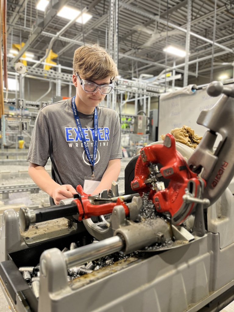 A person is operating a machine in an industrial workshop. They are wearing a grey T-shirt with 'EXETER WRESTLING' printed on it and a lanyard. The machine has red and metallic parts, with metal shavings visible, indicating active cutting or machining. The background includes shelves and various equipment in an organized workspace.