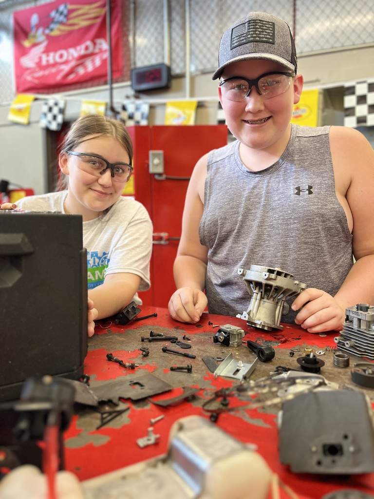 Two individuals are working on mechanical parts at a workbench in a workshop. The background features a red Honda banner, checkered flags, and various tools and equipment.