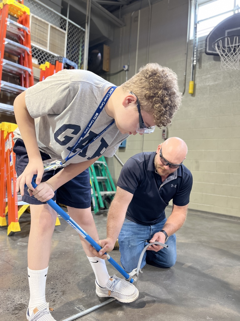 In a workshop, one person stands using a blue tool to bend a metal pipe while another kneels beside them to assist. The background includes ladders and various pieces of equipment.