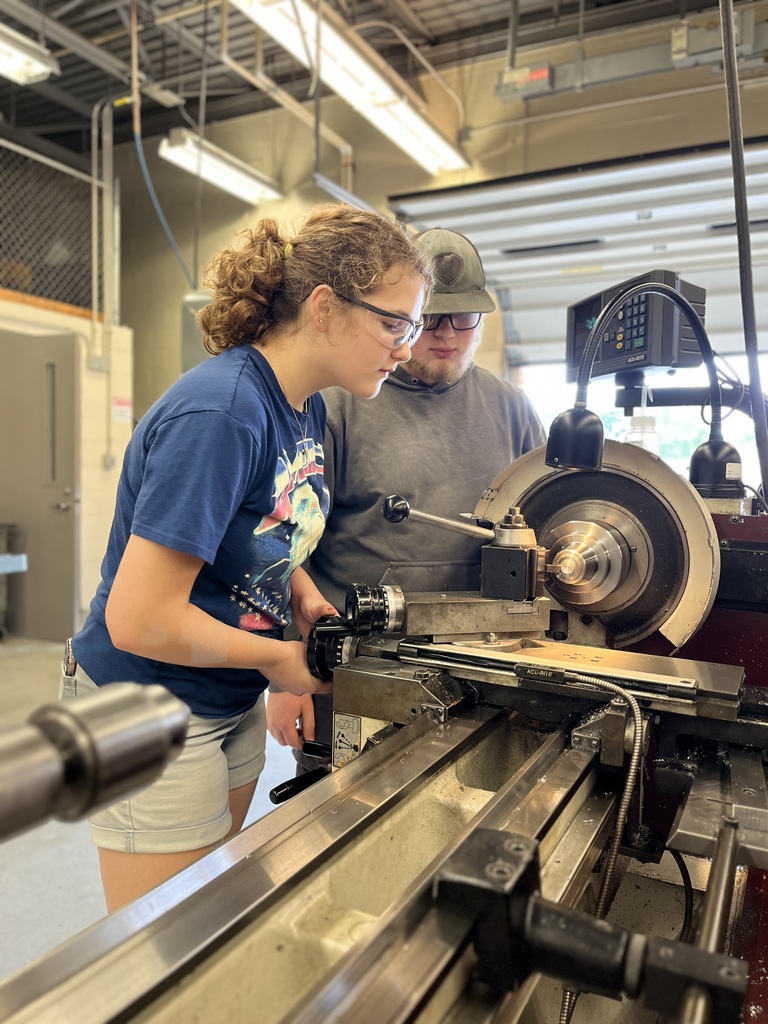 Two individuals are working with a lathe machine in an industrial workshop. The person in the foreground, wearing a blue T-shirt and shorts, is operating the machine, while another individual in a cap is partially visible. The lathe includes a rotating spindle, cutting tool, and a digital readout display.