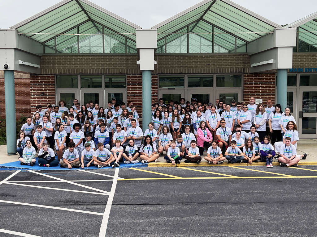 A large group of people, mostly young individuals, are gathered in front of a building with a glass roof and brick facade. They are all wearing matching light blue T-shirts and many have lanyards around their necks. The group is posing for a photo on the sidewalk and parking lot area in front of the building.