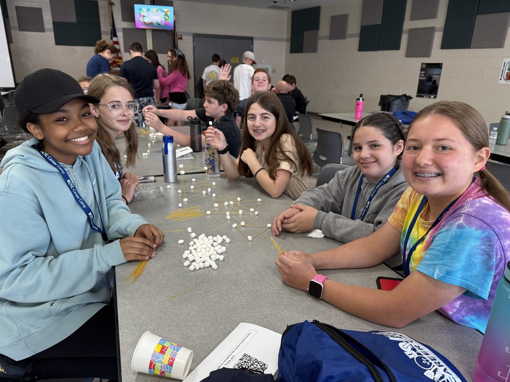 A group of students are seated around a table in a classroom or activity room, working on a project using uncooked spaghetti and marshmallows to build structures. Other student groups are visible in the background engaged in similar activities. The table holds water bottles, a cup with colorful designs, and some papers.